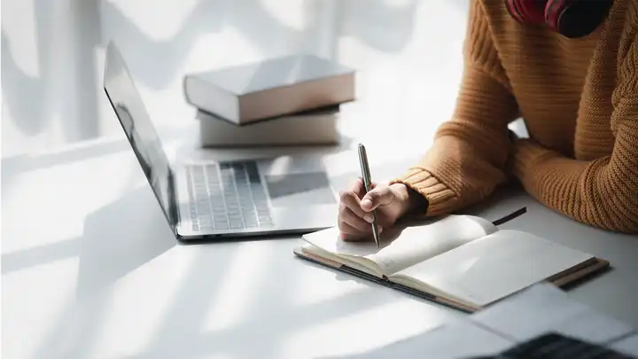 white wall and desk with hands taking notes next to laptop