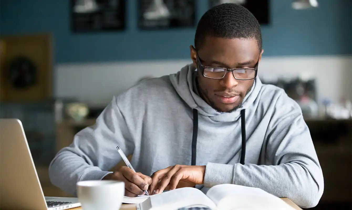 man writing notes from book at table with computer and coffee